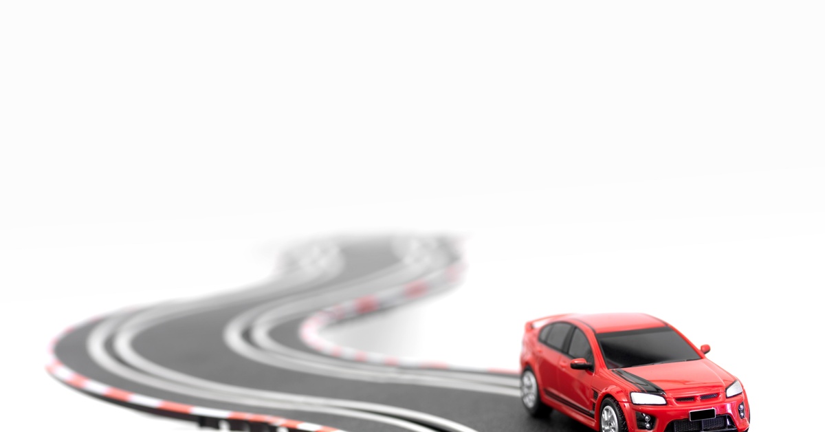 A red-and-black modern slot car in a sports car model on a two-rail track turning around a sharp corner.