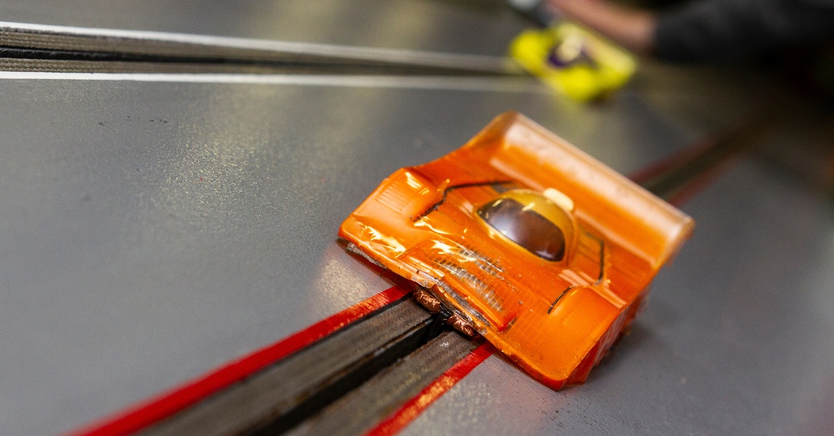 An orange slot car driving on a slot car track. There is a yellow slot car following behind it.