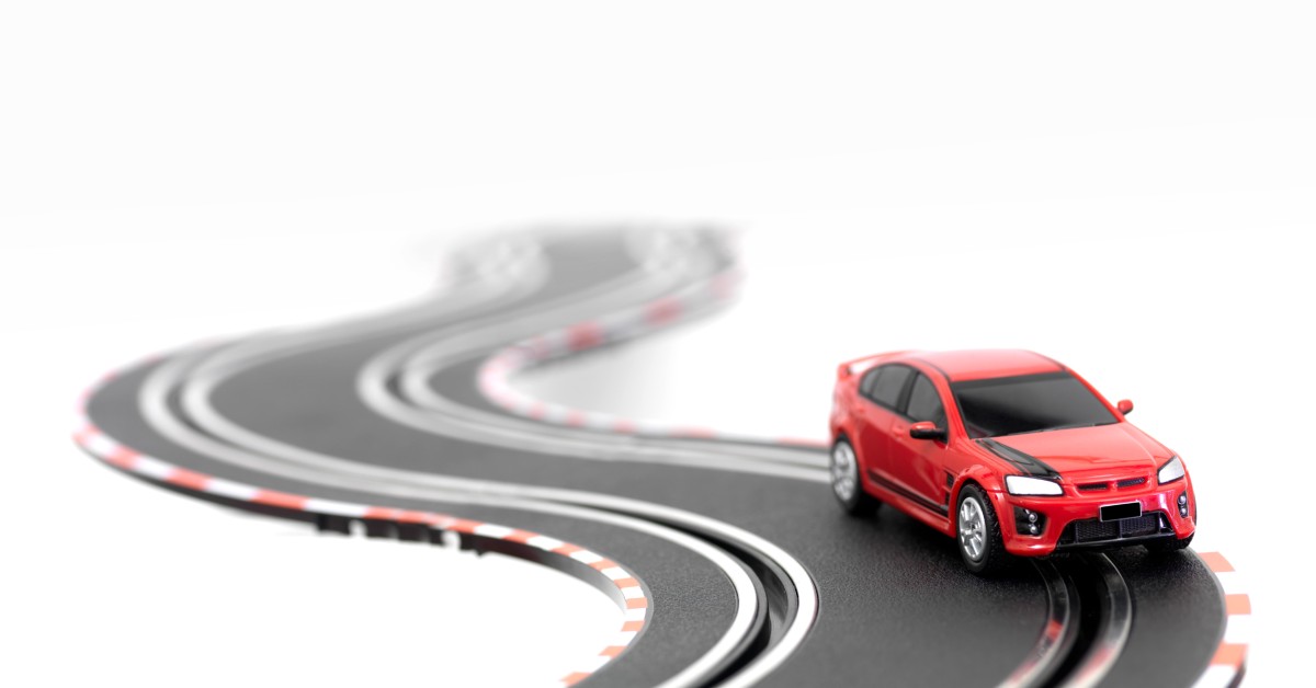 A sleek, red slot car situated on a length of race track. The race track disappears into the white background.
