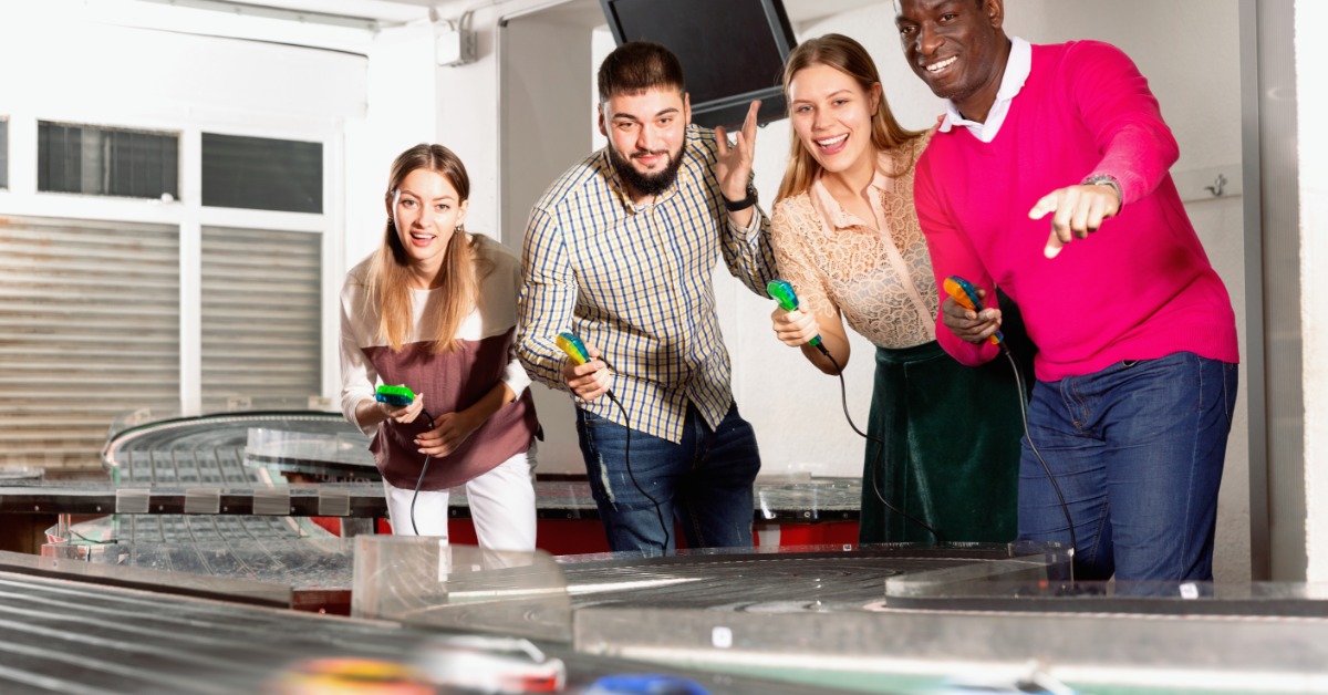 Two men and two women standing behind a slot car race track with sot car controllers in their hands.