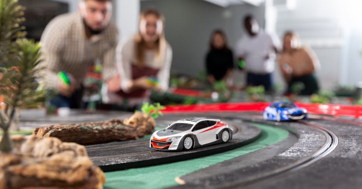 Two small slot cars racing on a track surrounded by foliage. Several people are standing behind the tracks.