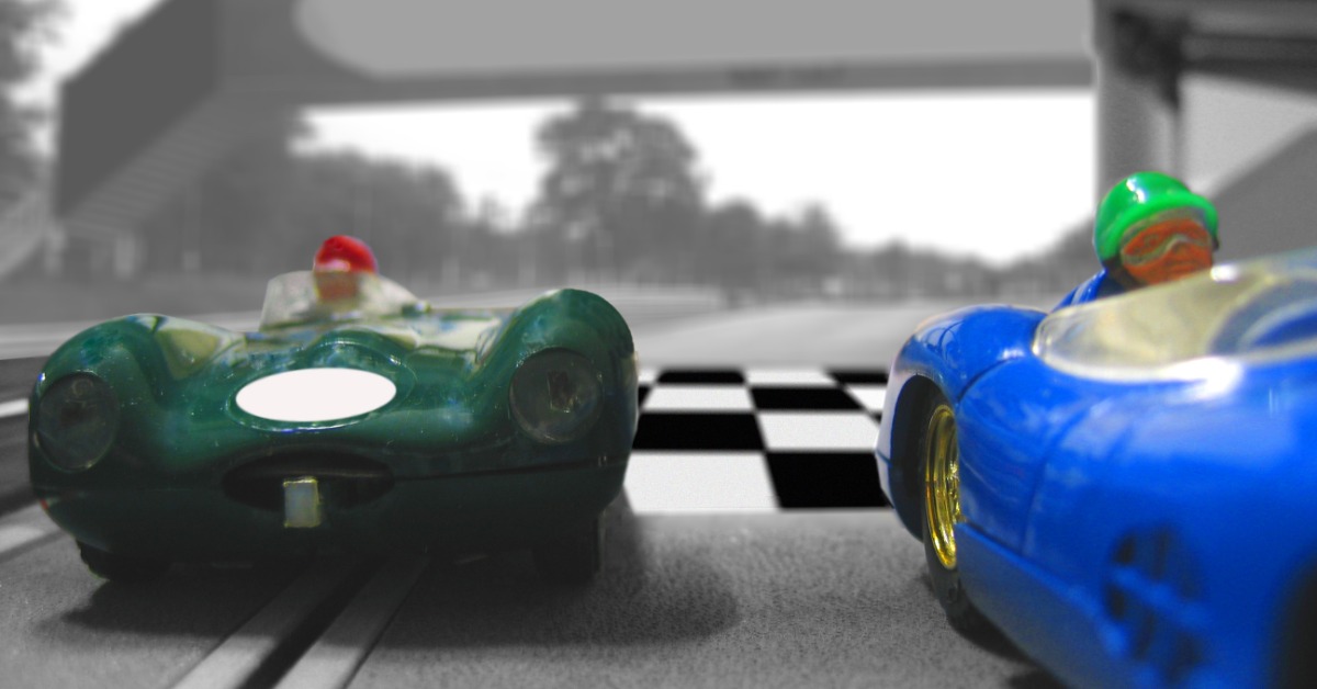 A close up on a pair of vintage slot cars, one green and the other blue. They are attached to a slot track.