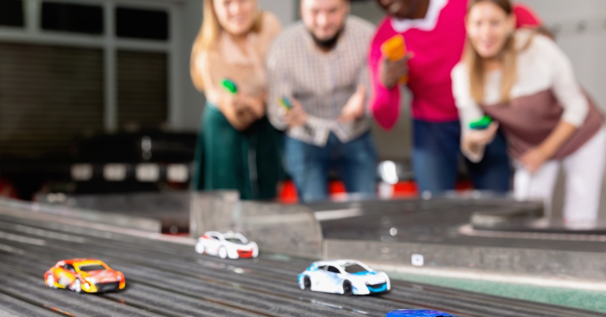 Four slot car racers in the background holding controllers for the slot cars they are racing on a wide track.