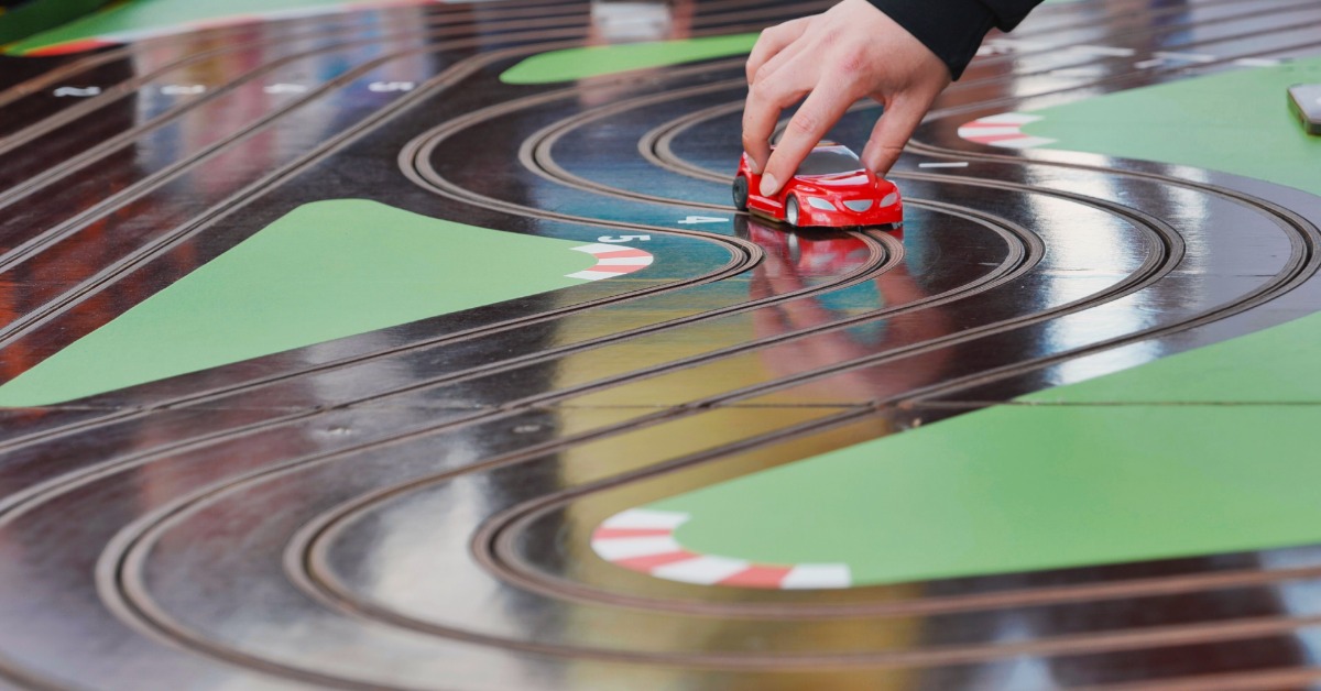 A person's hand carefully placing a red slot car down on to a slot car racing track that has multiple curves.