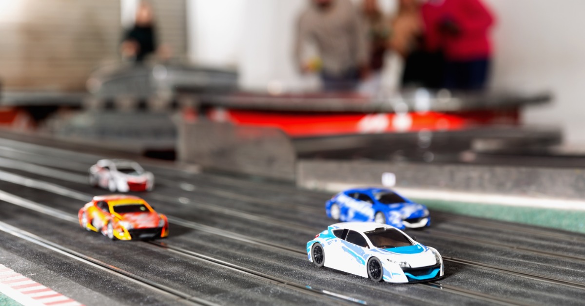 A closeup on four slot cars racing along a slot car track. There are people racing them blurred in the background.
