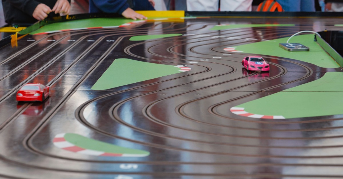 A large, winding slot car track with people playing with it. There are two cars on the track, one is red and one is pink.