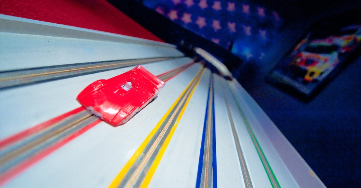 A plain red slot car sits in a red lane on a colorful slot-car racing track. Each lane is a different color.