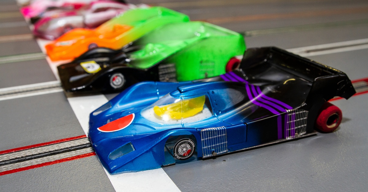 Slot cars sitting at the starting line. The front car is blue, purple, and black and all of the other cars are have various color combinations.
