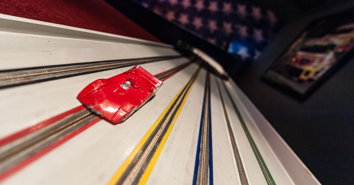 A red slot car speeding down a track with a blurry background and a poster of race cars to the right of it.