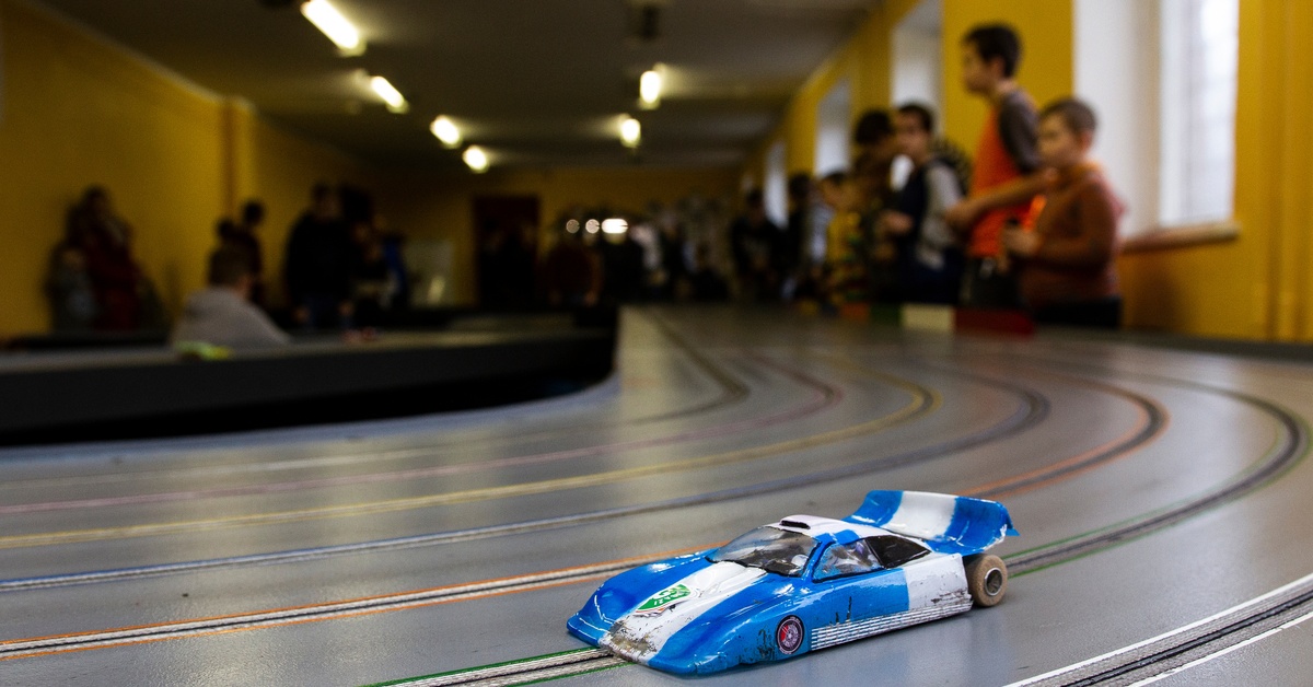 How To Succeed in Slot Car Racing Competitions A large slot car racing track with a blue and white slot car. Multiple people are standing around watching the race.