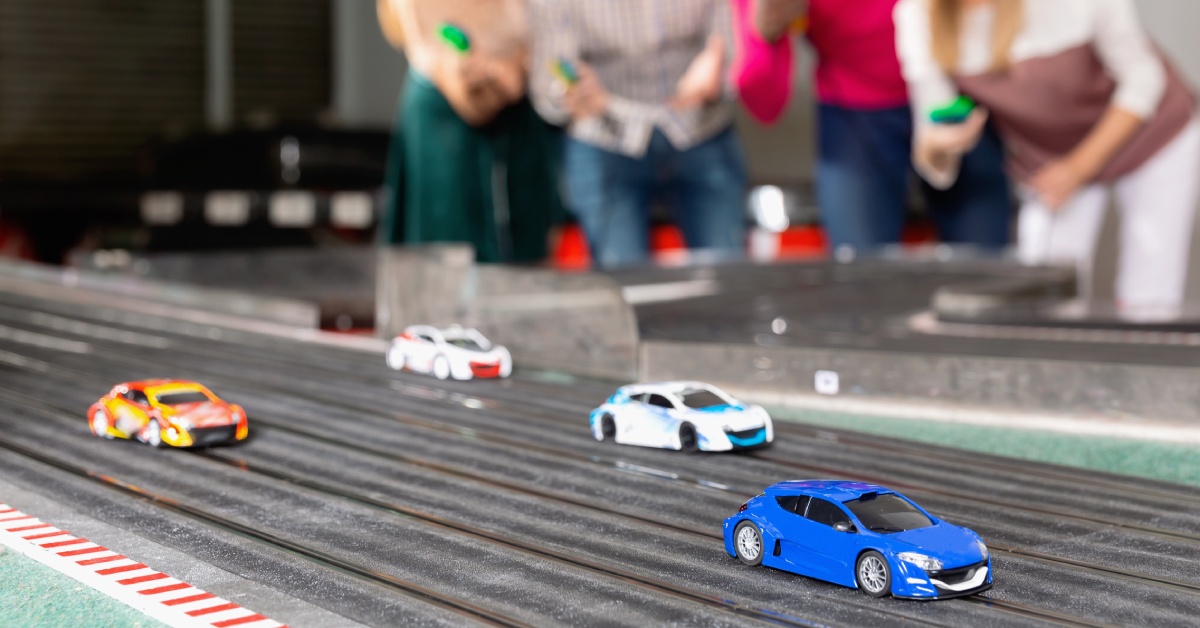 On a slot car track, there are multiple cars racing against each other while the racers hold the controllers behind them.