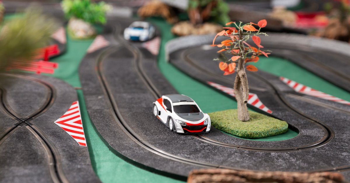 Two small slot cars drive around curves and straights, while passing small decorations, on a small slot car track.