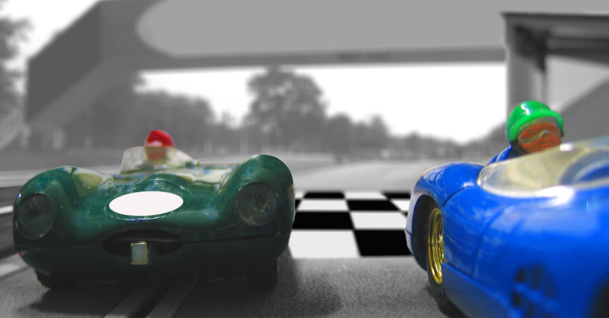 Two vintage slot cars square up at the beginning of a race track. The track has a black and white checkered finish line.
