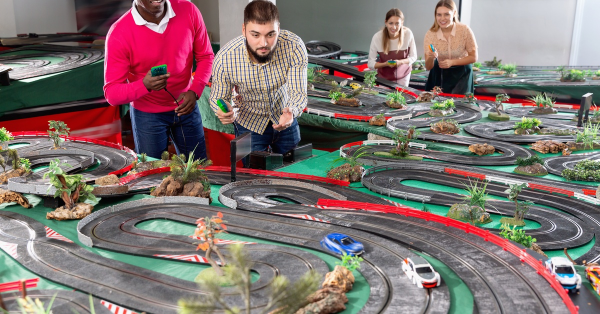Two men and two women gathered around a large and sprawling slot car race track watching the cars drive around.