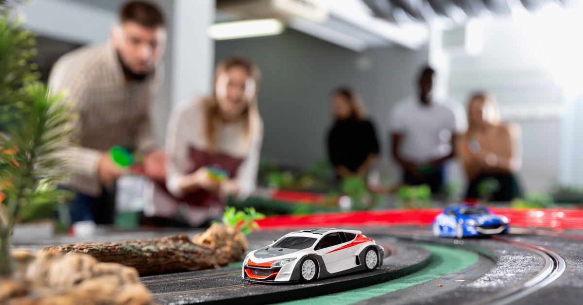 Two slot cars racing along a slot track with scenery pieces. There are several people in the background controlling the cars.