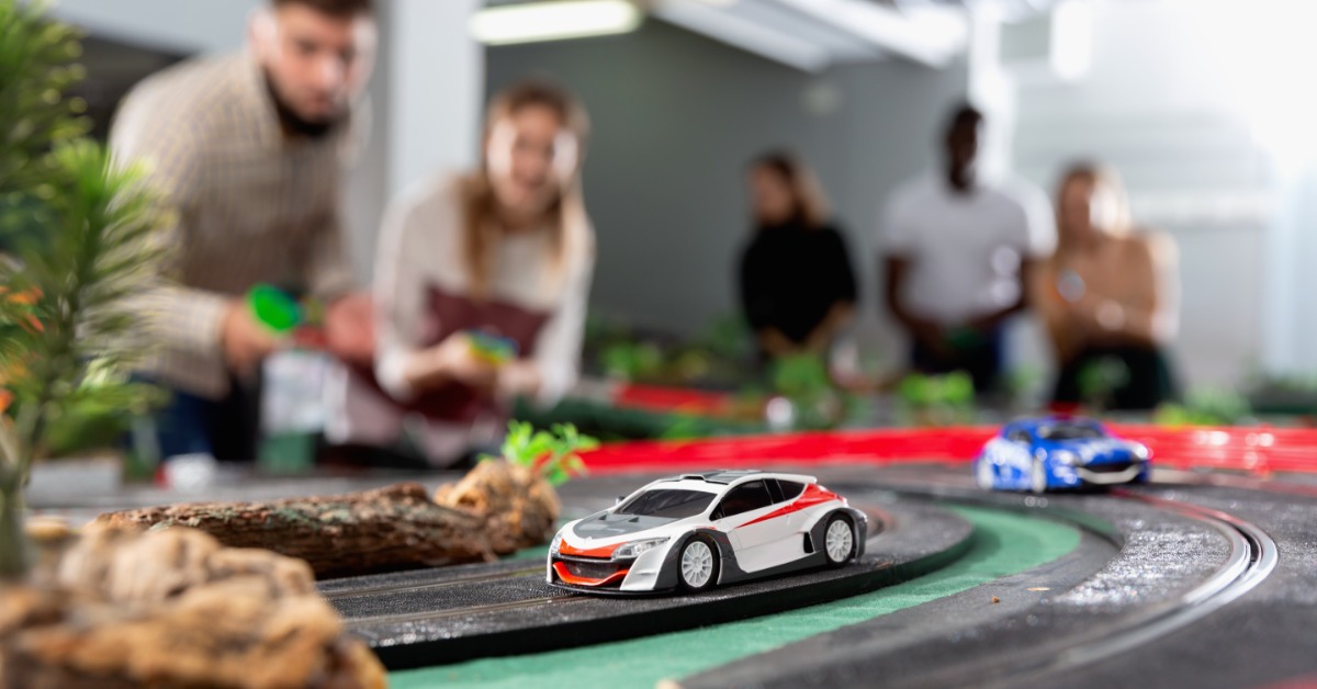 Two slot cars racing on a slot car track. There are multiple people controlling the cars in the background.