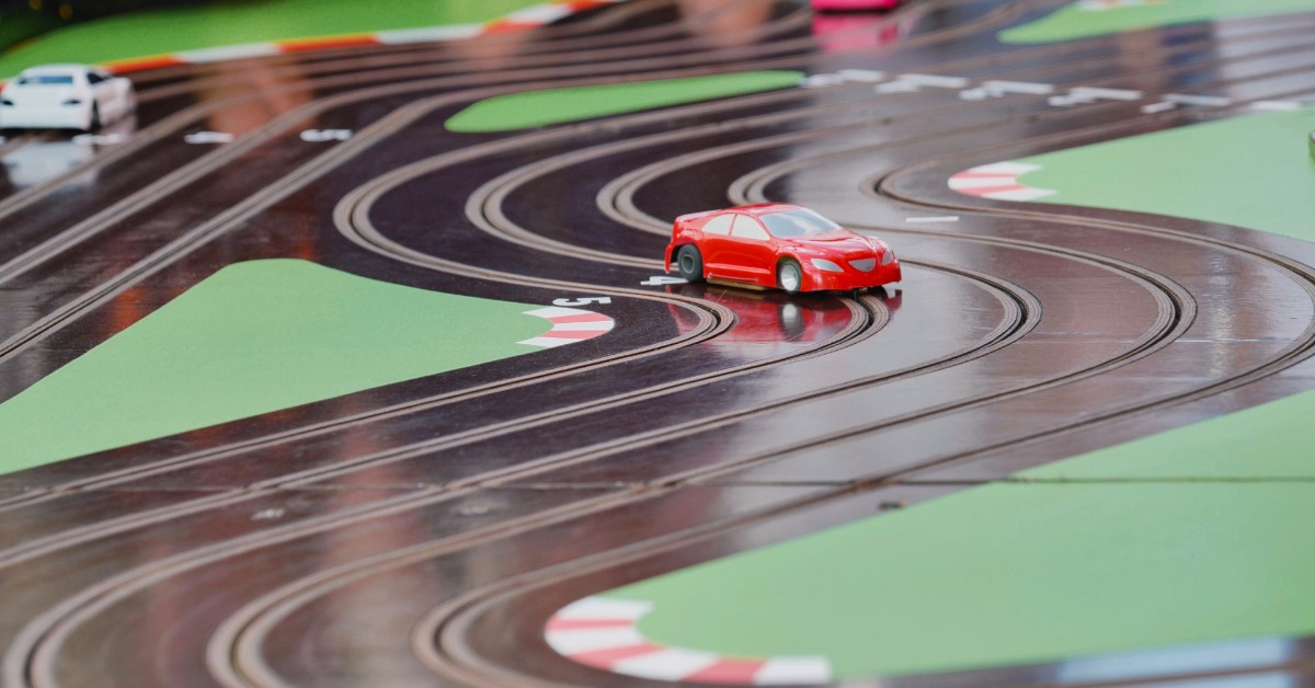 A small, red slot car coming around the corner of a slot car race track. Other slot cars are in the background.