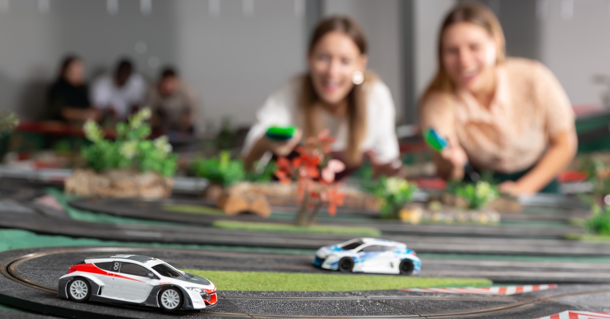 Two friends standing at the edge of a slot car racing track while they control and race two cars with their green controllers.