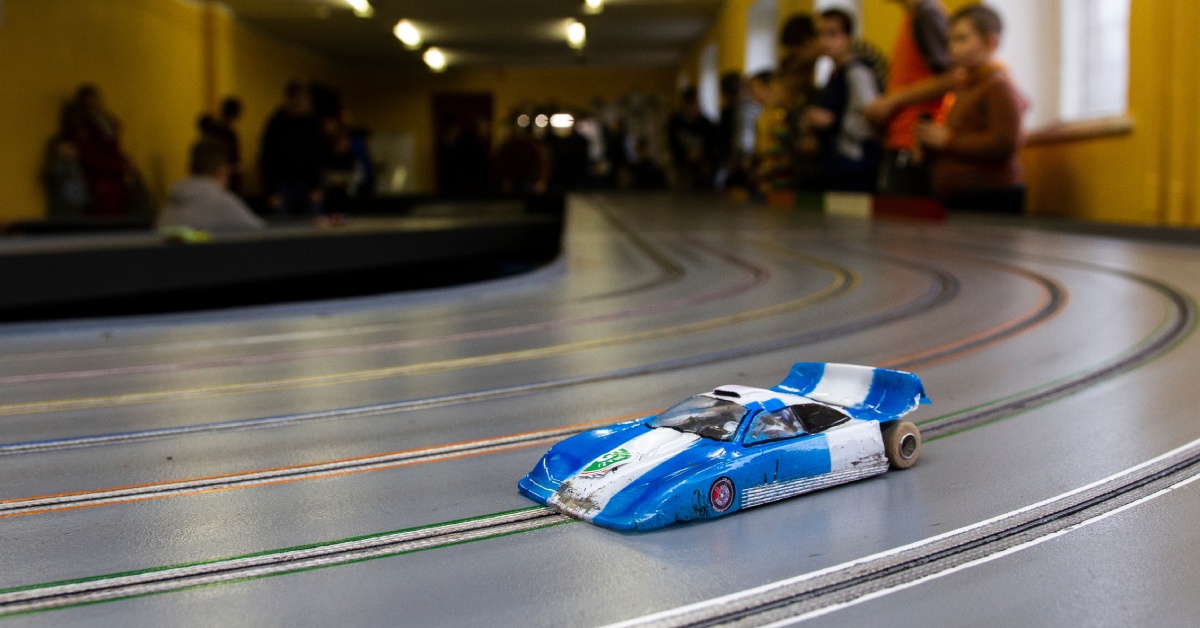 On a large slot car track, there is a small blue slot car racing down the track while viewers watch in the distance.