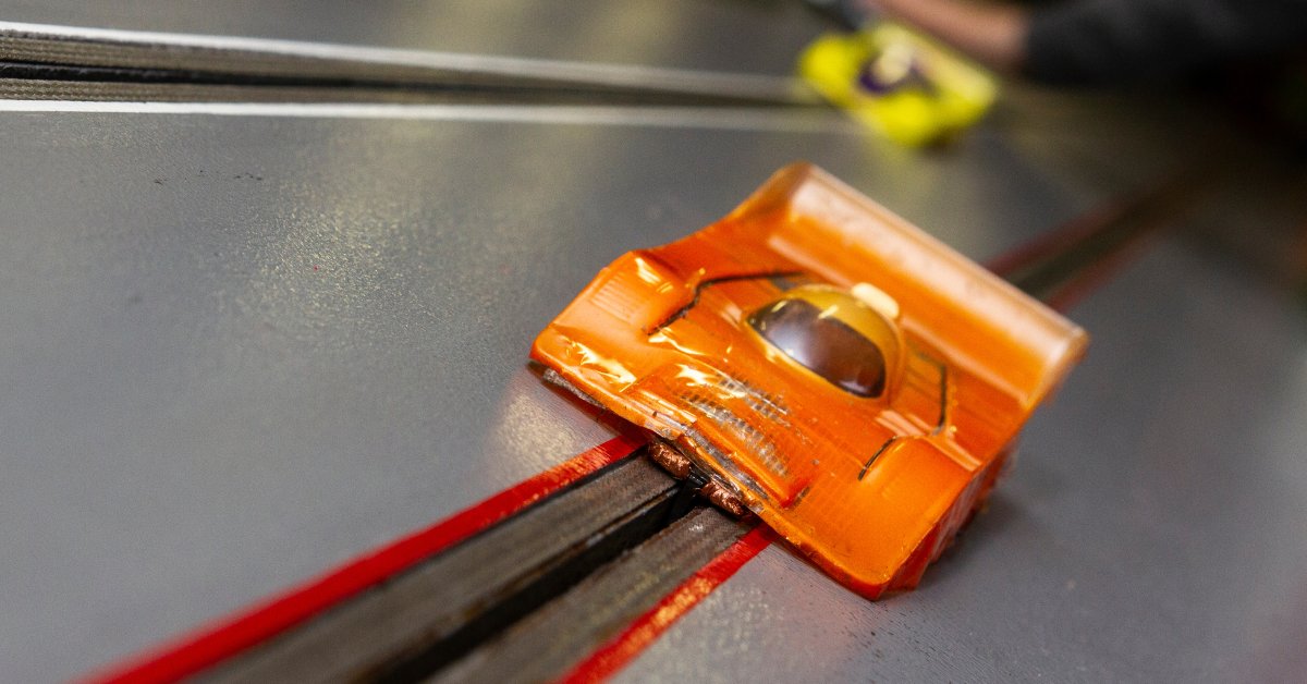 A close up of an orange slot car racing on a small track with a blurry yellow slot car in the distance behind it.